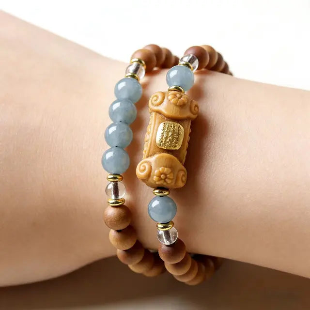 Hand wearing a Tibetan-style beaded bracelet with natural wood beads, light blue jade stones, and a carved centerpiece, minimalist background