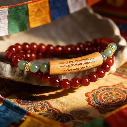 Handmade red agate beaded bracelet with green jade-like accents and a carved wooden tube, displayed on a textured fabric with Tibetan patterns and colorful prayer flags in soft focus.