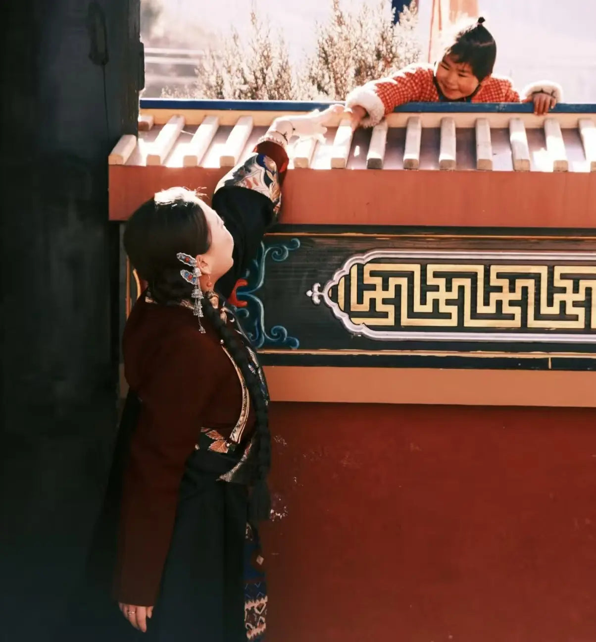 A Tibetan girl in traditional clothing stands on tiptoes, reaching out to touch a young child standing against a red wall.