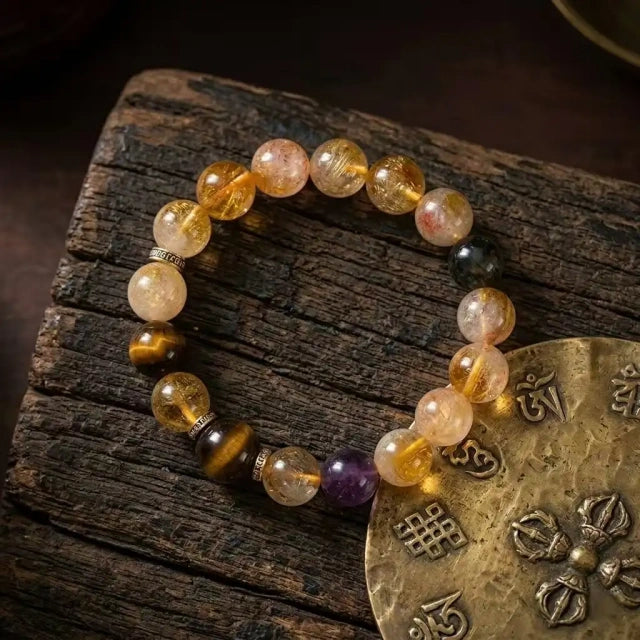 A feng shui multi-gemstone healing bracelet laid flat on a rustic wooden surface next to a traditional brass Tibetan Buddhist amulet disk.