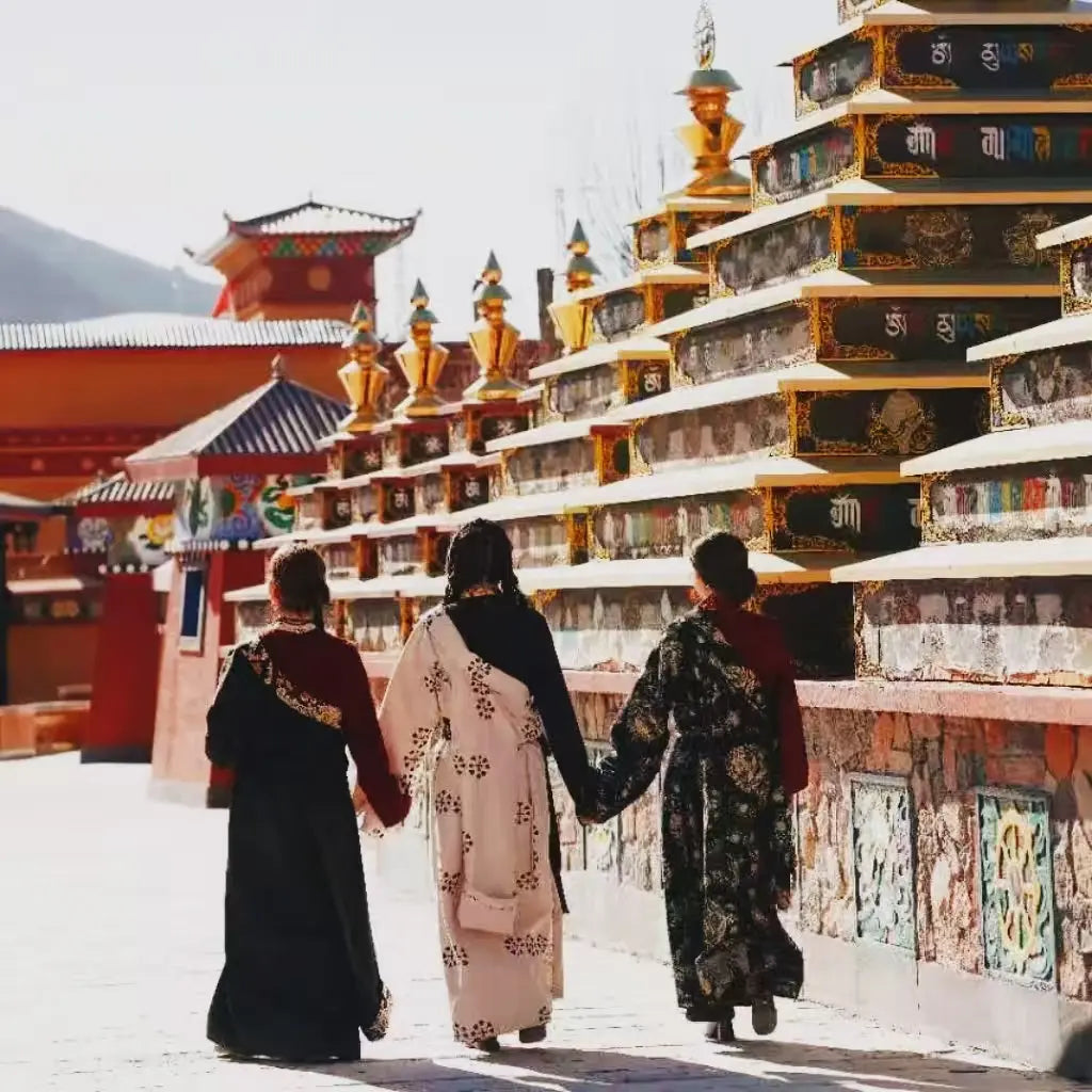 Three women wearing traditional Tibetan dress chuba holding hands while walking past a row of prayer wheels at a Buddhist temple.